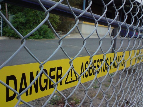 Yellow tape reads 'Danger! Asbestos Hazard' behind chain link fence